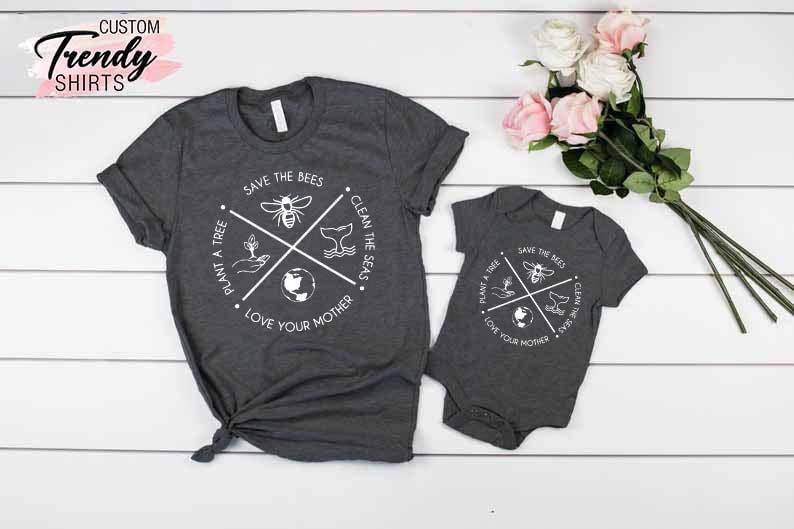 Op de afbeelding: Een set van twee grijze t-shirts met een wit grafisch ontwerp. Het grotere shirt heeft een rond ontwerp met de tekst "Save the Bees", "Plant a Tree", "Clean the Seas" en "Love Your Mother". Het kleinere shirt is een baby-romper met hetzelfde ontwerp.