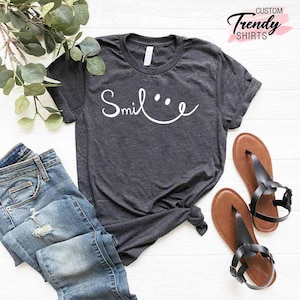 Op de afbeelding: Een donkergrijs T-shirt met de tekst "Smile..." in witte letters. Het shirt is geknoopt aan de voorkant en wordt gecombineerd met een blauwe jeans en zwarte sandalen.