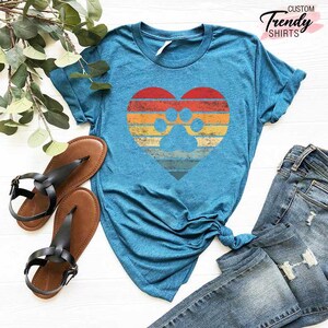 Op de afbeelding: Een tealkleurig T-shirt met een hartvormig ontwerp met een pootafdruk. Het hart heeft een retro zonsondergang kleurenschema van rode, oranje en gele strepen. De afbeelding toont ook bruine sandalen en een blauwe jeans.