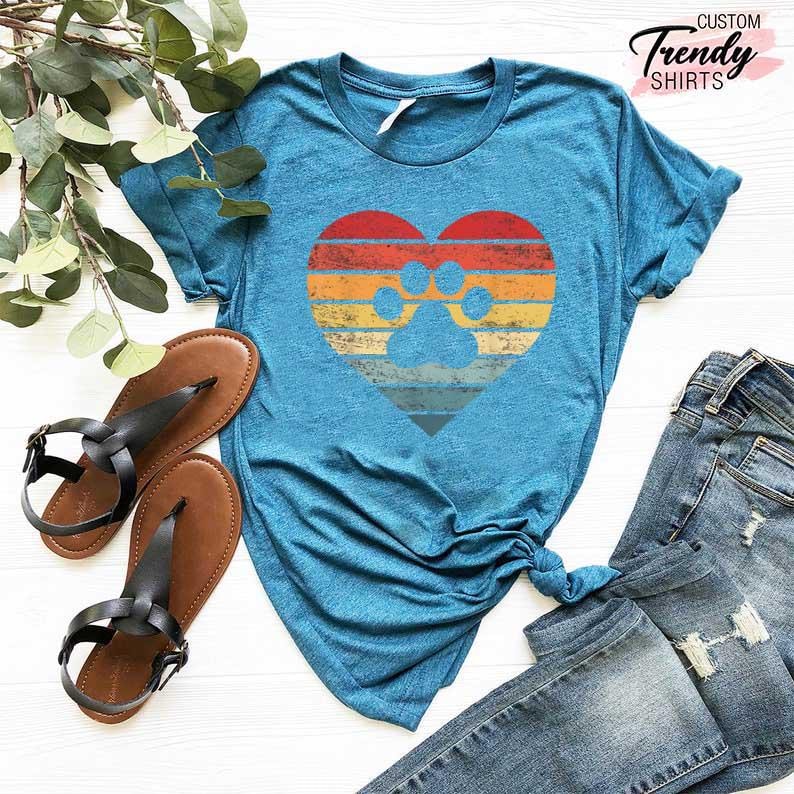 Op de afbeelding: Een tealkleurig T-shirt met een hartvormig ontwerp met een pootafdruk. Het hart heeft een retro zonsondergang kleurenschema van rode, oranje en gele strepen. De afbeelding toont ook bruine sandalen en een blauwe jeans.