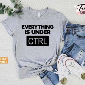 K&ouml;nnte beinhalten: Ein hellgraues T-Shirt mit dem Text "EVERYTHING IS UNDER CTRL" in Schwarz. Das Shirt wird mit einer blauen Jeans und wei&szlig;en Turnschuhen kombiniert. Das Bild enth&auml;lt auch einen Zweig mit gr&uuml;nen Bl&auml;ttern.
