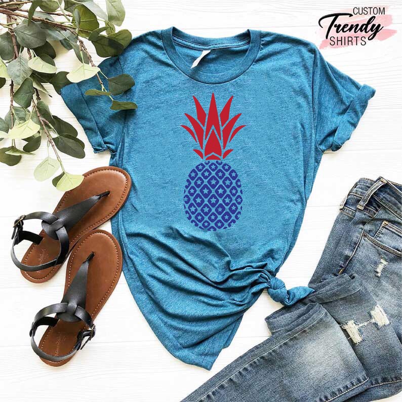 May include: A teal blue t-shirt with a red and blue pineapple design. The pineapple is patterned with white stars and blue diamonds.