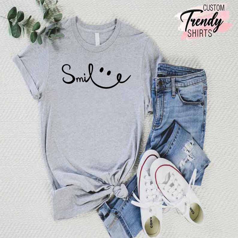 Op de afbeelding: Een grijs T-shirt met de tekst "Smile..." in zwarte letters. Het shirt is gestyled met een blauwe jeans en witte sneakers.
