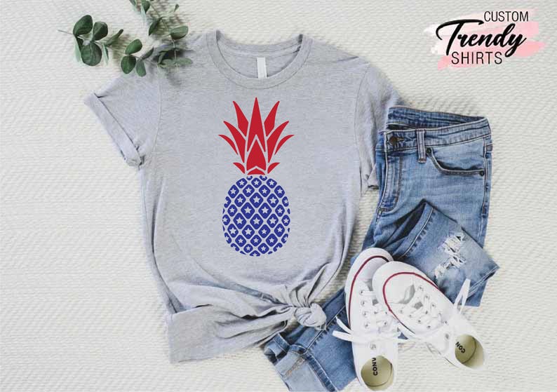 May include: A gray t-shirt with a red and blue pineapple design. The pineapple is patterned with white stars and blue diamonds. The shirt is laid out on a white surface with a pair of blue jeans and white sneakers.