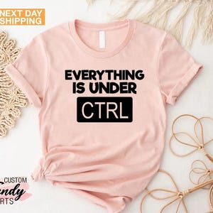 K&ouml;nnte beinhalten: Hellrosa T-Shirt mit dem schwarzen Text "EVERYTHING IS UNDER CTRL". Das Wort "CTRL" befindet sich in einem schwarzen Rechteck. Das Shirt hat einen Rundhalsausschnitt und kurze &Auml;rmel. Der Schriftzug "NEXT DAY SHIPPING" befindet sich in einer gelben Box.