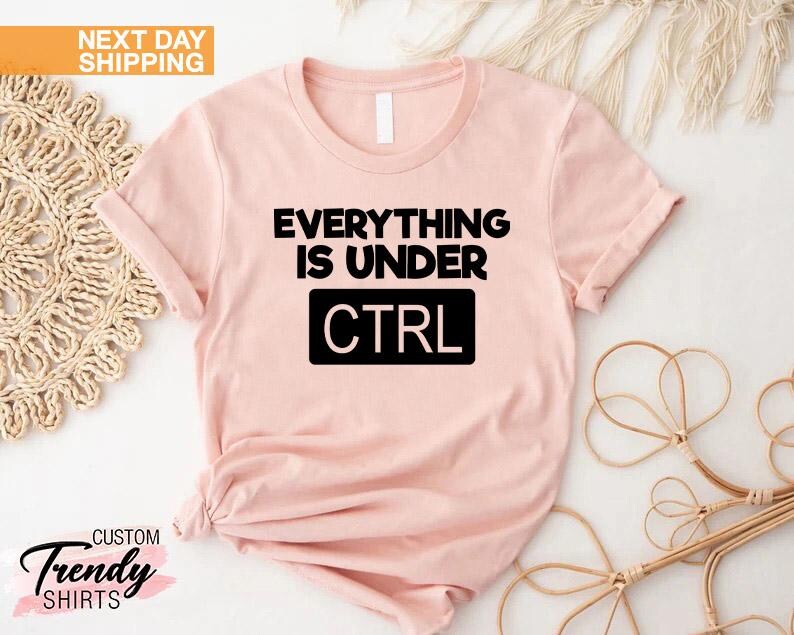 K&ouml;nnte beinhalten: Hellrosa T-Shirt mit dem schwarzen Text "EVERYTHING IS UNDER CTRL". Das Wort "CTRL" befindet sich in einem schwarzen Rechteck. Das Shirt hat einen Rundhalsausschnitt und kurze &Auml;rmel. Der Schriftzug "NEXT DAY SHIPPING" befindet sich in einer gelben Box.