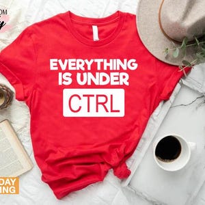 K&ouml;nnte beinhalten: Ein rotes T-Shirt mit dem wei&szlig;en Text "EVERYTHING IS UNDER CTRL". Das Shirt wird mit einem Hut, einem Tablett mit einer Tasse Kaffee, einer Uhr und einem Buch pr&auml;sentiert.
