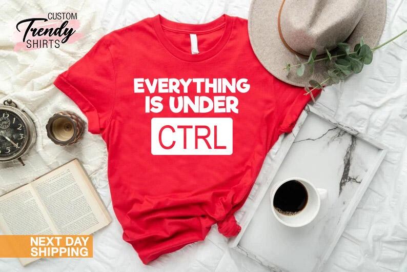 K&ouml;nnte beinhalten: Ein rotes T-Shirt mit dem wei&szlig;en Text "EVERYTHING IS UNDER CTRL". Das Shirt wird mit einem Hut, einem Tablett mit einer Tasse Kaffee, einer Uhr und einem Buch pr&auml;sentiert.