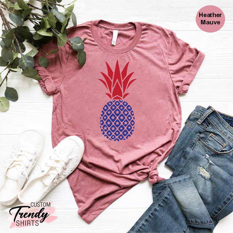May include: A heather mauve t-shirt with a red and blue pineapple design. The pineapple has a red top and a blue body with white stars. The shirt is laid flat on a white surface with a pair of white sneakers and blue jeans.