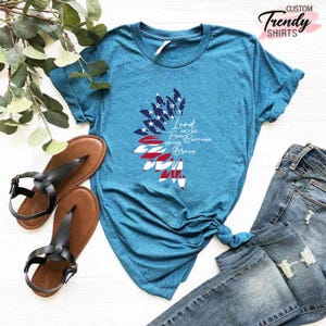 May include: A teal blue t-shirt with a white graphic of an American flag in the shape of a sunflower. The text "Land of the Free Because of the Brave" is printed on the shirt.