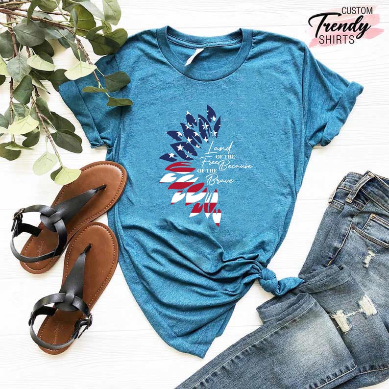 May include: A teal blue t-shirt with a white graphic of an American flag in the shape of a sunflower. The text "Land of the Free Because of the Brave" is printed on the shirt.