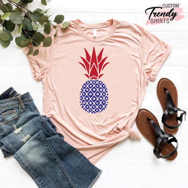 May include: A pink t-shirt with a red and blue pineapple design. The pineapple is patterned with white stars and blue diamonds. The shirt is on a white wooden surface.