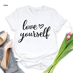 May include: White t-shirt with the words "love yourself" in a black script font, with a heart symbol above the word "yourself". The shirt is displayed with a pair of beige high heels, blue jeans, and a bouquet of pink tulips.
