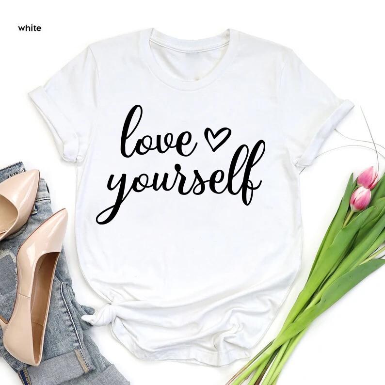 May include: White t-shirt with the words "love yourself" in a black script font, with a heart symbol above the word "yourself". The shirt is displayed with a pair of beige high heels, blue jeans, and a bouquet of pink tulips.