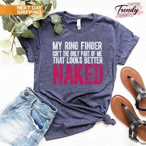May include: A navy blue t-shirt with the text "MY RING FINGER ISN'T THE ONLY PART OF ME THAT LOOKS BETTER NAKED" in white and pink. The shirt is paired with denim shorts and brown sandals.