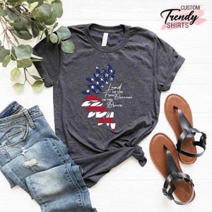 May include: A dark gray t-shirt with a red, white, and blue American flag design. The text "Land of the Free Because of the Brave" is printed on the shirt.