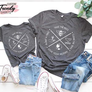 Op de afbeelding: Twee grijze T-shirts met een wit grafisch ontwerp. De grafiek toont een cirkel met een kruis erin. Het kruis heeft vier symbolen: een bij, een boom, een walvis en een wereldbol. De tekst rond de cirkel luidt "Plant een boom", "Red de bijen", "Maak de zee&euml;n schoon" en "Liefheb je moeder".