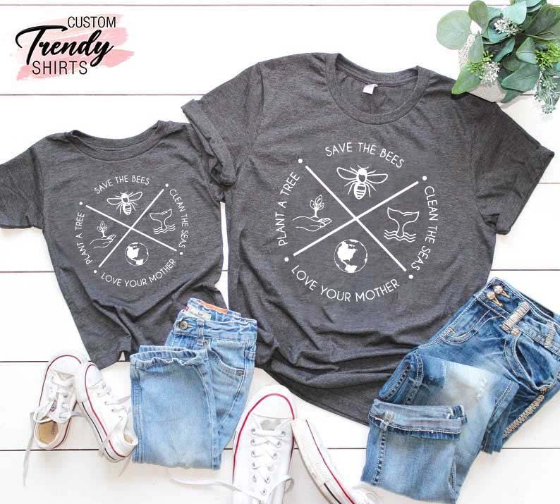 Op de afbeelding: Twee grijze T-shirts met een wit grafisch ontwerp. De grafiek toont een cirkel met een kruis erin. Het kruis heeft vier symbolen: een bij, een boom, een walvis en een wereldbol. De tekst rond de cirkel luidt "Plant een boom", "Red de bijen", "Maak de zee&euml;n schoon" en "Liefheb je moeder".