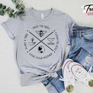 Op de afbeelding: Grijs T-shirt met een zwart grafisch ontwerp met een kruis met vier symbolen: een bij, een boom, een walvis en de aarde. De tekst rond het kruis luidt "Save the Bees", "Plant a Tree", "Clean the Seas" en "Love Your Mother".