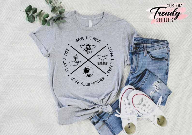 Op de afbeelding: Grijs T-shirt met een zwart grafisch ontwerp met een kruis met vier symbolen: een bij, een boom, een walvis en de aarde. De tekst rond het kruis luidt "Save the Bees", "Plant a Tree", "Clean the Seas" en "Love Your Mother".