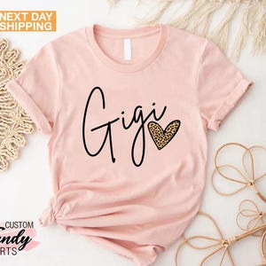May include: A pink t-shirt with the word "Gigi" written in black cursive script and a heart with a leopard print design.
