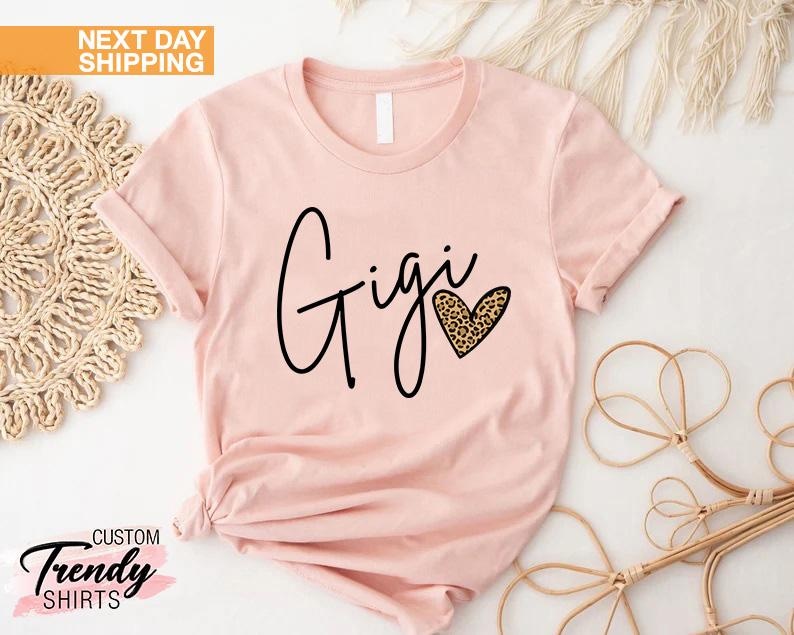 May include: A pink t-shirt with the word "Gigi" written in black cursive script and a heart with a leopard print design.
