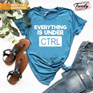 K&ouml;nnte beinhalten: Ein t&uuml;rkisfarbenes T-Shirt mit dem wei&szlig;en Text "EVERYTHING IS UNDER CTRL". Das Shirt wird mit braunen Sandalen mit schwarzen Riemen und einer blauen Jeans kombiniert. Das Bild enth&auml;lt auch einen Zweig mit Gr&uuml;n.