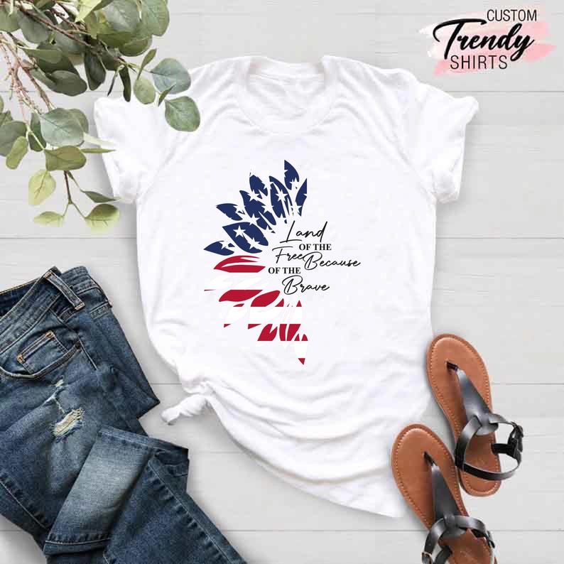 May include: White t-shirt with a red, white, and blue sunflower design. The sunflower is made up of stars and stripes. The text "Land of the Free Because of the Brave" is printed on the shirt.