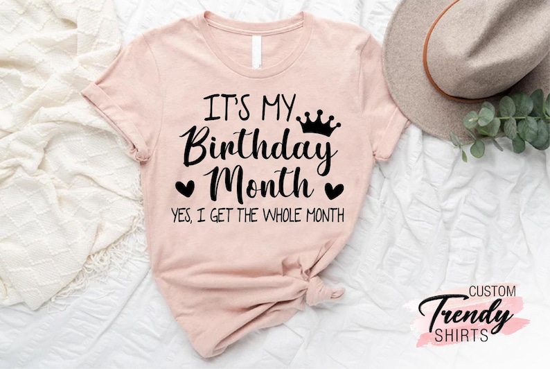 May include: A pink t-shirt with the text "It's my Birthday Month. Yes, I get the whole month." in black lettering. A crown graphic is above the word "Birthday".