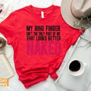 May include: A red t-shirt with the text "MY RING FINGER ISN'T THE ONLY PART OF ME THAT LOOKS BETTER NAKED" in black and pink. The shirt is displayed with a hat, a tray with a coffee cup, a book, and a clock.