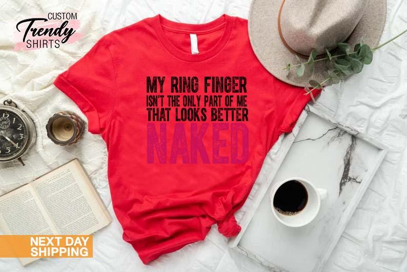 May include: A red t-shirt with the text "MY RING FINGER ISN'T THE ONLY PART OF ME THAT LOOKS BETTER NAKED" in black and pink. The shirt is displayed with a hat, a tray with a coffee cup, a book, and a clock.