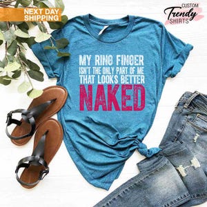 May include: A turquoise t-shirt with the text "MY RING FINGER ISN'T THE ONLY PART OF ME THAT LOOKS BETTER NAKED" in white and pink. The shirt is paired with blue jeans and brown sandals with black straps. The image also includes a sprig of greenery.