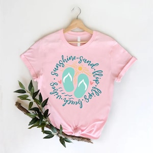 K&ouml;nnte beinhalten: Rosa T-Shirt mit einem Grafikdesign von Flip-Flops, einer Sonne und dem Text "sunshine. sand. flip. flop. beach. vibes."