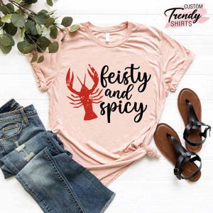 May include: A pink t-shirt with a red crawfish and the text "feisty and spicy" in black.
