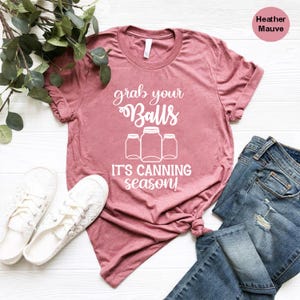 May include: Heather mauve t-shirt with the text "grab your Balls, it's canning season!" in white. The shirt is styled with white sneakers and blue jeans. The shirt has a crew neck and short sleeves.