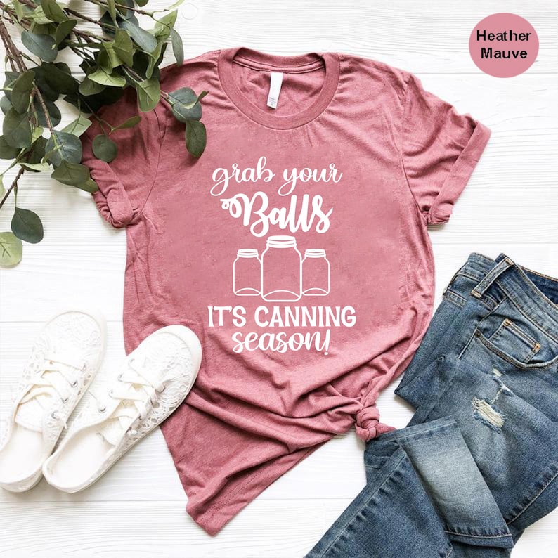 May include: Heather mauve t-shirt with the text "grab your Balls, it's canning season!" in white. The shirt is styled with white sneakers and blue jeans. The shirt has a crew neck and short sleeves.