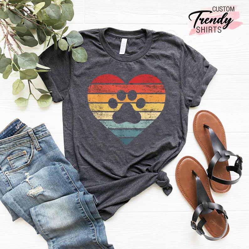 Op de afbeelding: Donkergrijs t-shirt met een retro hartontwerp en een pootafdruk. Het hart heeft een gestreept patroon in rood, oranje, geel en blauw. De tekst "Custom Trendy Shirts" staat rechtsboven. Het shirt wordt gecombineerd met een blauwe jeans en bruine sandalen.