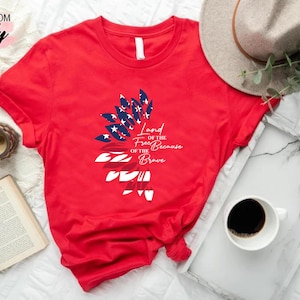 May include: Red t-shirt with a graphic of an American flag in the shape of a feather. The text "Land of the Free Because of the Brave" is printed on the shirt.