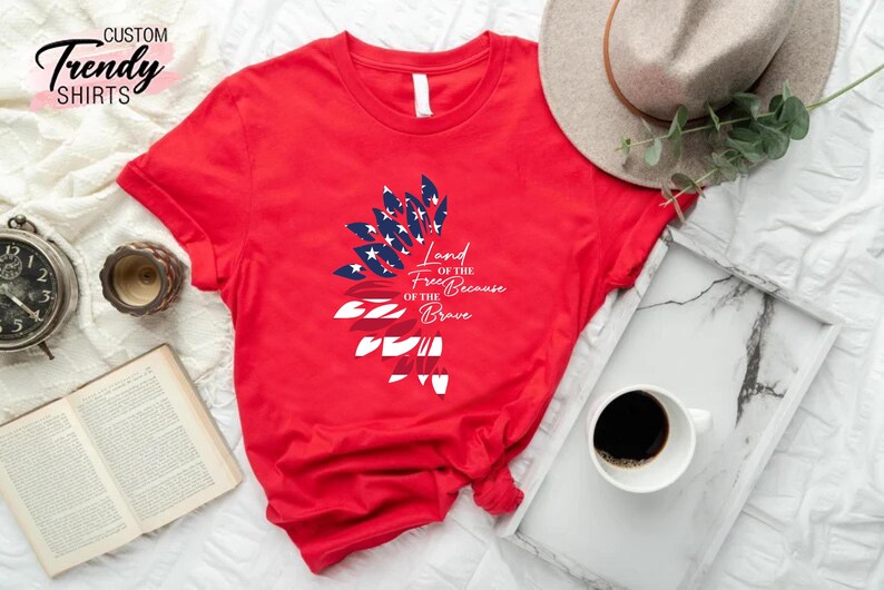 May include: Red t-shirt with a graphic of an American flag in the shape of a feather. The text "Land of the Free Because of the Brave" is printed on the shirt.