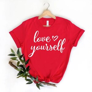 May include: A red t-shirt with the words "love yourself" in white script. The shirt is hanging on a wooden hanger. A sprig of greenery and berries is placed below the shirt.