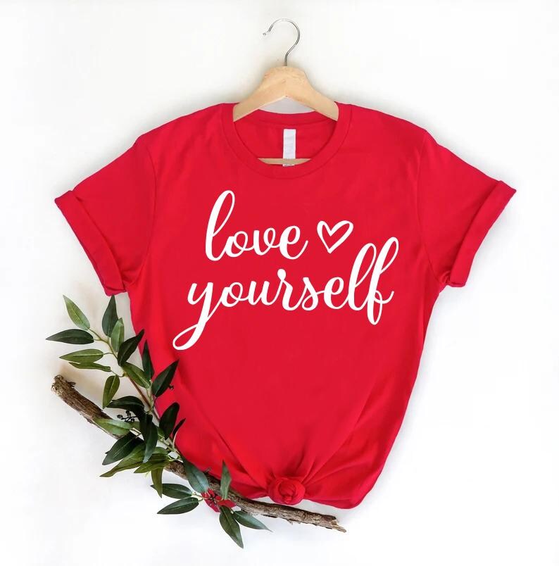 May include: A red t-shirt with the words "love yourself" in white script. The shirt is hanging on a wooden hanger. A sprig of greenery and berries is placed below the shirt.
