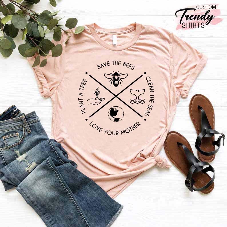 Op de afbeelding: Een roze T-shirt met een zwart grafisch ontwerp met de tekst "Red de bijen, plant een boom, maak de zee&euml;n schoon, hou van je moeder". De grafiek is een cirkel met een kruis in het midden. Het kruis toont een bij, een boom, een walvis en een wereldbol.