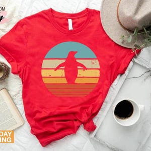 May include: Red t-shirt with a retro-style graphic of a penguin standing in front of a sunset. The graphic is in shades of blue, yellow, orange, and red.