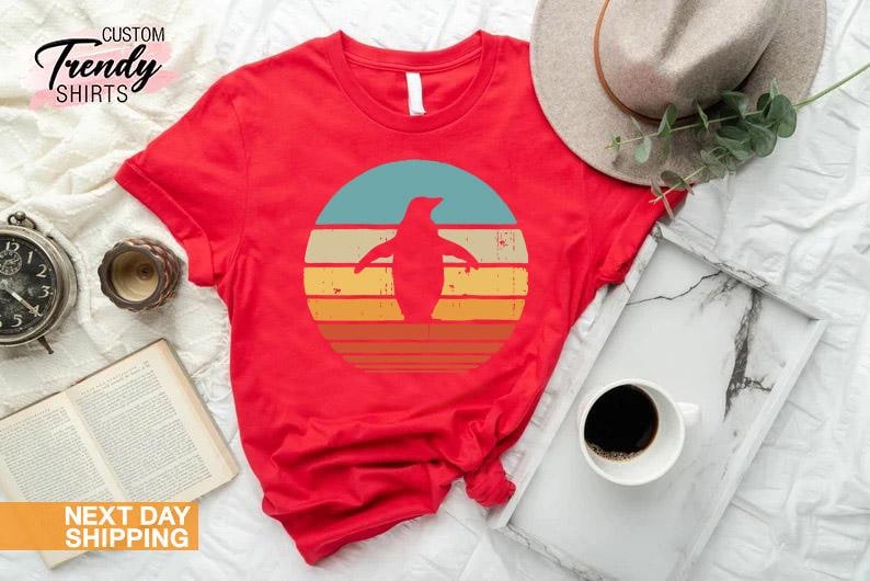 May include: Red t-shirt with a retro-style graphic of a penguin standing in front of a sunset. The graphic is in shades of blue, yellow, orange, and red.