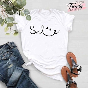 Op de afbeelding: Wit T-shirt met zwarte tekst die "Smile" zegt met een smiley ernaast getekend. Het shirt ligt op een houten oppervlak met een paar bruine sandalen en een blauwe jeans.