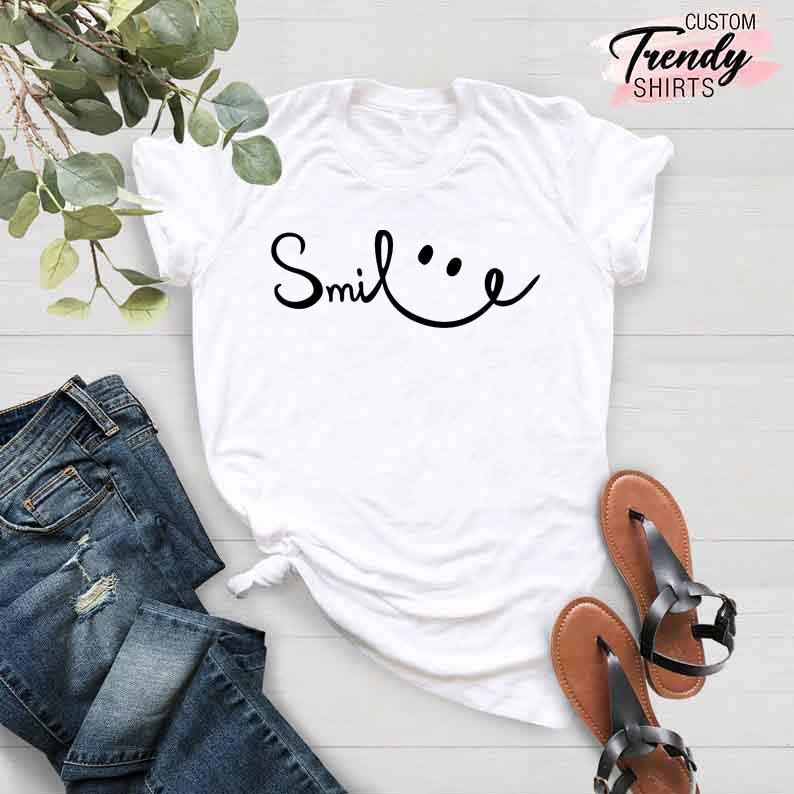 Op de afbeelding: Wit T-shirt met zwarte tekst die "Smile" zegt met een smiley ernaast getekend. Het shirt ligt op een houten oppervlak met een paar bruine sandalen en een blauwe jeans.