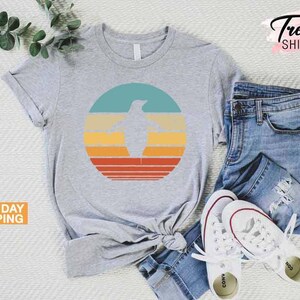 May include: A gray t-shirt with a retro-style graphic of a penguin in a circle with orange, yellow, and blue stripes. The shirt is laid out on a white surface with a pair of blue jeans and white sneakers.