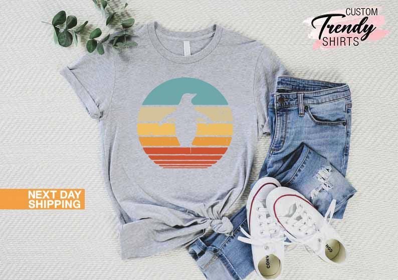 May include: A gray t-shirt with a retro-style graphic of a penguin in a circle with orange, yellow, and blue stripes. The shirt is laid out on a white surface with a pair of blue jeans and white sneakers.