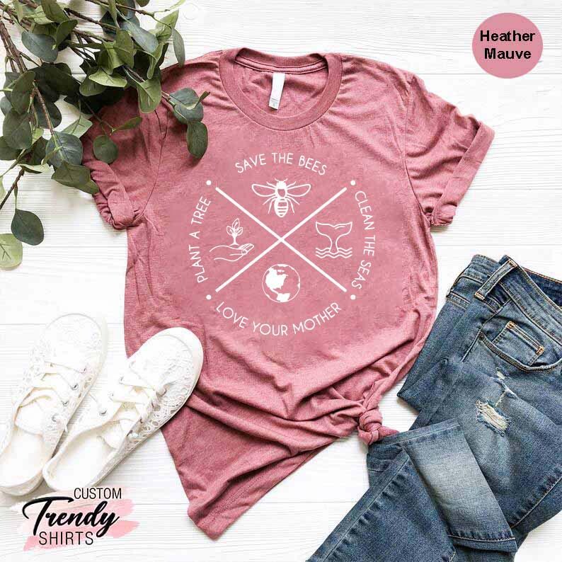 Op de afbeelding: Een heather mauve t-shirt met een wit grafisch ontwerp met een kruis met vier symbolen: een bij, een walvisstaart, een wereldbol en een hand die een plant vasthoudt. De tekst rond de symbolen luidt: "Save the Bees", "Clean the Seas", "Plant a Tree" en "Love Your Mother".