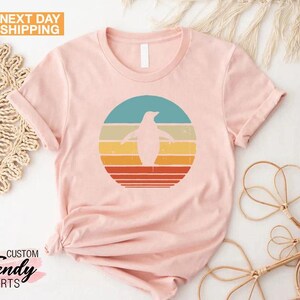 May include: A pink t-shirt with a retro-style graphic of a penguin standing in front of a sunset. The graphic is in shades of blue, yellow, orange, and red.